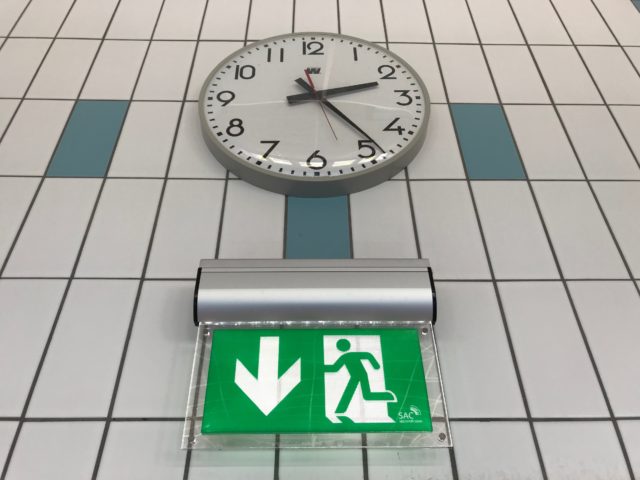 Clock On Tiled Wall With Emergency Sign Underneath It