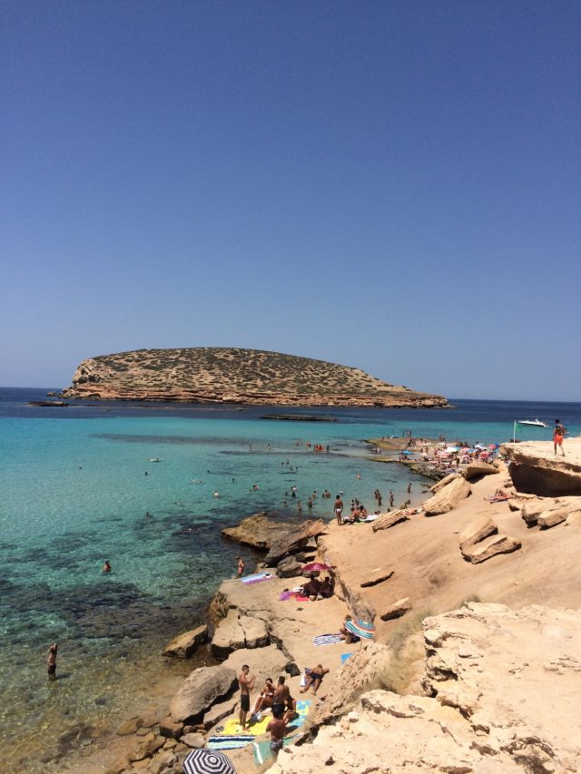 Beach With Sunbathers And Swimmers With An Island In Ibiza