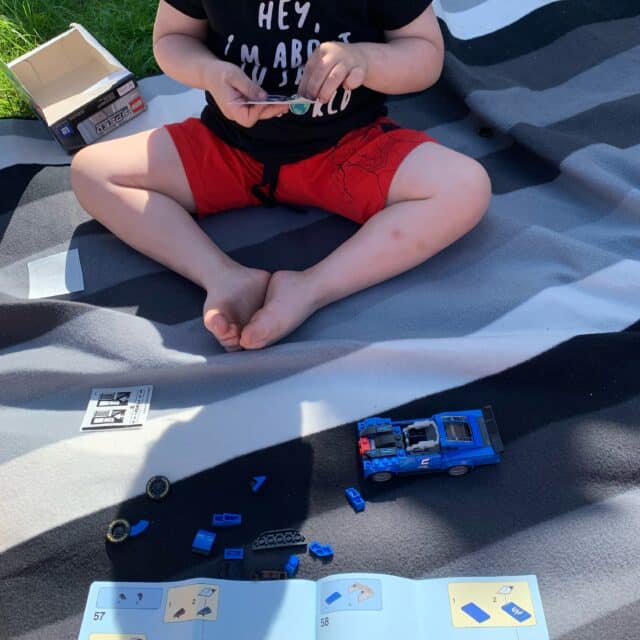 Boy Sits On A Picnic Blanket And Builds A Lego Car