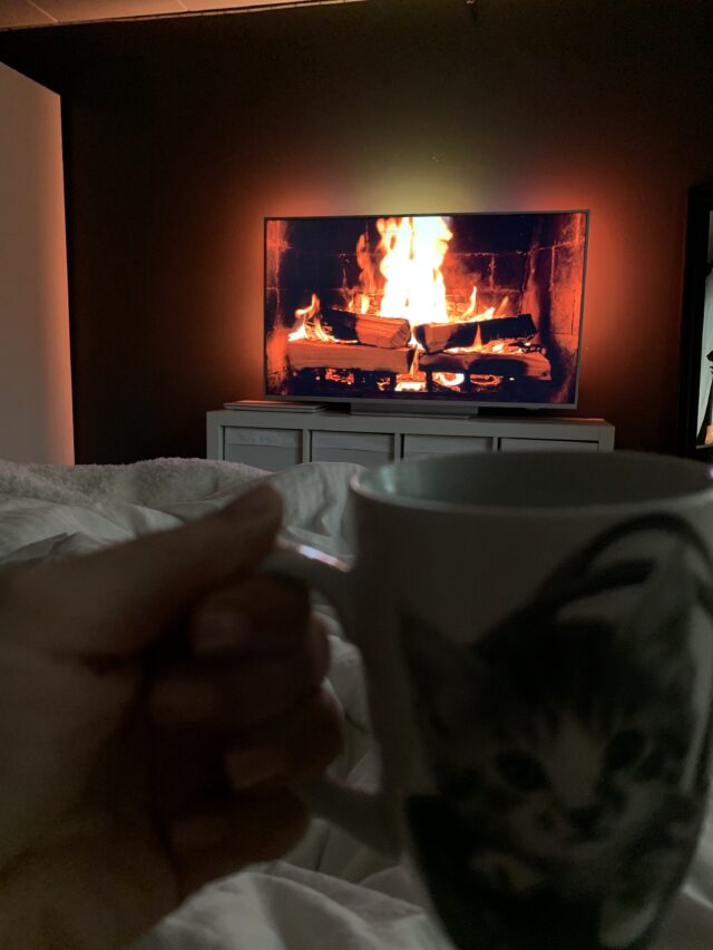 Person In Bed Holding Coffee Mug In Hand Watching Fireplace